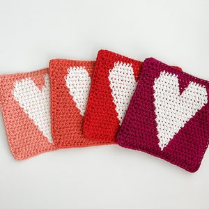 May include: Four crocheted square washcloths in shades of pink, coral, red, and burgundy. Each cloth features a white heart design in the center. The washcloths are arranged side by side on a white surface.