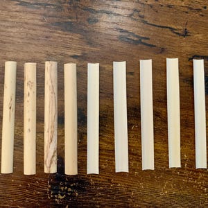 May include: Ten natural, unpainted, wooden drinking straws of varying lengths and widths. The straws are arranged in a row on a wooden surface.