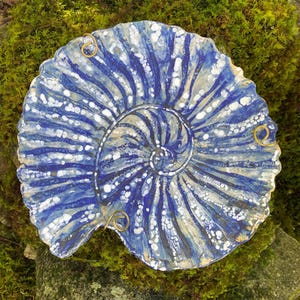 May include: A circular ceramic plate featuring a blue and white spiral pattern resembling an ammonite fossil. The surface has a textured, mottled glaze with radiating lines. Three small gold-toned metal wire loops are attached to the scalloped edge.