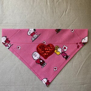 May include: Pink fabric dog bandana with a red sequined heart and a pattern of cartoon characters from the Peanuts comic strip. The characters are holding hearts and some are wearing hats.