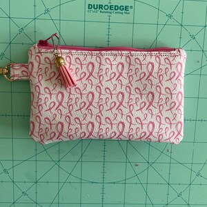 May include: A white zippered pouch with a pink ribbon pattern and a pink tassel. The pouch is on a green cutting mat with markings in inches.