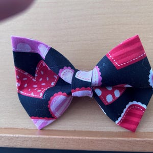 May include: A fabric bow tie with a black base, decorated with red and pink heart and polka dot designs. The bow tie is a classic shape and is suitable for pets or people. The bow tie is handmade.
