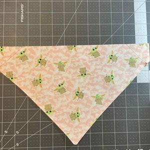 May include: Pink fabric triangle bandana with a repeating pattern of green baby Yoda characters.