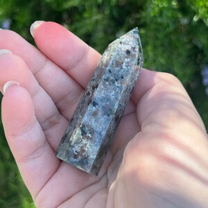 May include: A green and black speckled crystal point, also known as a wand, is held in a hand. The crystal is smooth and polished.