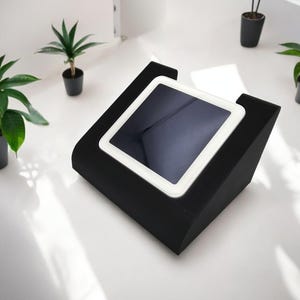 May include: A black tablet stand with a white tablet on top. The stand is angled upwards and has a soft, padded surface. The tablet has a black screen and a white border.