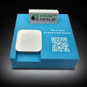 May include: A blue countertop display with a white square-shaped card reader and a business card with the text "Print Realm". The display also features a QR code with the text "Tap or Scan To Follow Our Socials".
