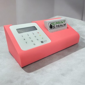 May include: A pink plastic stand with a white card reader and a business card holder. The card reader has a small screen and a keypad with numbers 0-9, a tick, and a cross. The business card holder has a white card with the text "PRINT REALM" and a green and black logo.