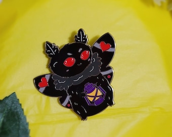 May include: Black enamel pin of a cartoon moth with red eyes, holding a purple lantern. The moth has a fluffy body and red hearts on its wings.