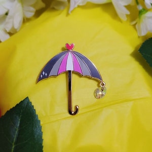 May include: A gold-tone pin in the shape of an umbrella with a pink, white, and gray striped canopy. The umbrella has a small heart at the top and a dangling crystal at the bottom.