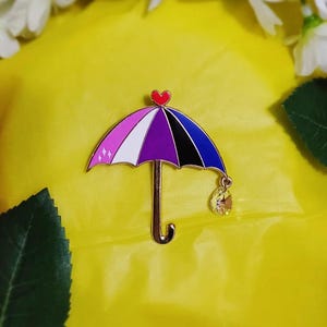 May include: A colorful umbrella pin with a heart on top and a dangling crystal charm. The umbrella features pink, white, purple, black, and blue sections. The pin is set against a bright yellow background.