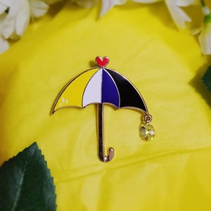 May include: A gold-tone enamel pin shaped like an umbrella with a yellow, white, blue, and black design. The umbrella has a red heart at the top and a green crystal hanging from the bottom.