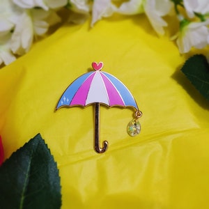 May include: A pink, blue, and white enamel pin shaped like an umbrella with a gold heart at the top and a gold chain with a clear crystal hanging from the bottom.