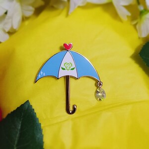 May include: A blue and white enamel pin shaped like an umbrella with a green rose design and a red heart at the top. A clear crystal charm hangs from the bottom of the umbrella.
