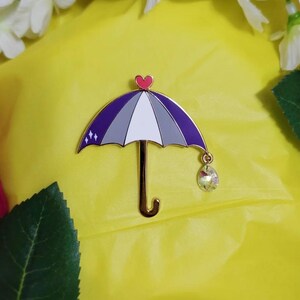 May include: A decorative umbrella pin with a gold-colored handle and a purple, gray, and white canopy. The pin features a small red heart at the top and a crystal charm hanging from the bottom. The pin is set against a yellow background.