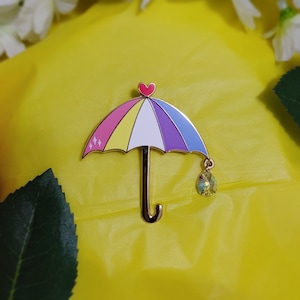 May include: A rainbow-colored enamel pin shaped like an umbrella with a heart at the top and a dangling crystal.