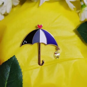 May include: A pin shaped like an umbrella with a gold handle and a heart at the top. The umbrella's canopy is divided into black, gray, white, and blue sections. A crystal charm hangs from the umbrella. The pin is set against a yellow background.