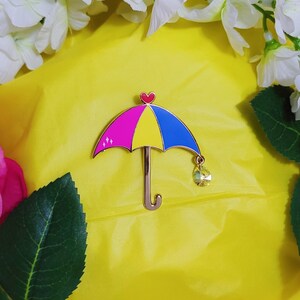 May include: A colorful umbrella pin with pink, yellow, and blue sections. The pin has a gold-colored stem and a heart at the top. A teardrop-shaped crystal hangs from the umbrella.
