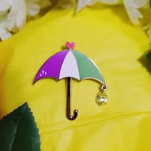 May include: A colorful umbrella-shaped enamel pin with a gold-tone finish. The umbrella features purple, white, and green panels, a small pink heart at the top, and a yellow crystal charm. The pin is set against a bright yellow background.