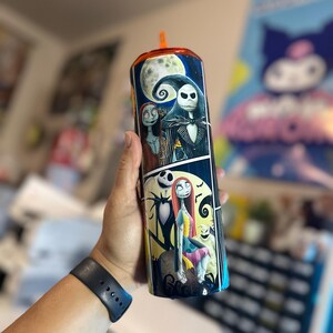 May include: A black and white stainless steel tumbler with a colorful image of Jack Skellington and Sally from the Nightmare Before Christmas. The tumbler has a black lid with a clear straw.