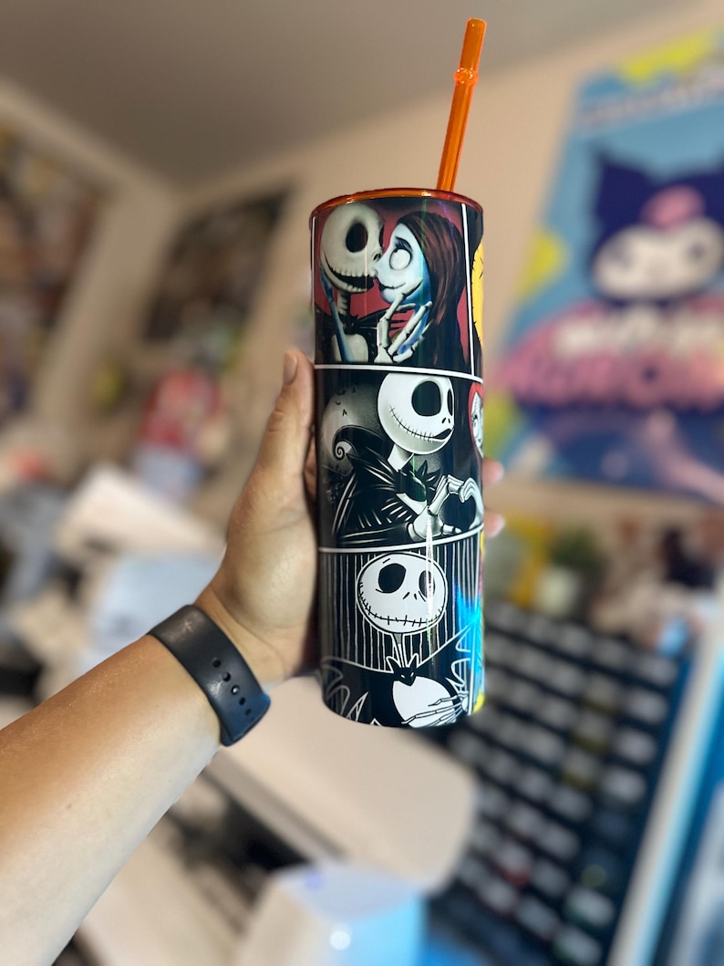 May include: A black and white tumbler with a graphic of Jack Skellington and Sally from the Nightmare Before Christmas. The tumbler has a black lid and an orange straw.