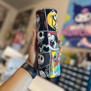 May include: A black and white tumbler with a comic book style design featuring characters from The Nightmare Before Christmas. The characters include Jack Skellington, Sally, and Oogie Boogie.