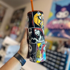 May include: A black and white stainless steel tumbler with a comic book style design featuring characters from The Nightmare Before Christmas. The tumbler has an orange straw and is filled with a clear liquid.