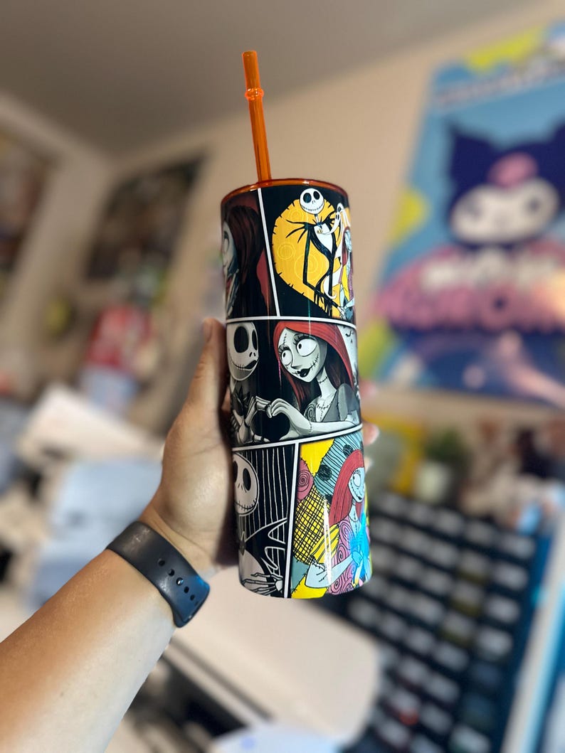 May include: A black and white stainless steel tumbler with a comic book style design featuring characters from The Nightmare Before Christmas. The tumbler has an orange straw and is filled with a clear liquid.