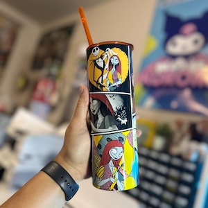 May include: A black and white tumbler with a colorful comic book style design featuring Jack Skellington and Sally from The Nightmare Before Christmas. The tumbler has an orange straw.