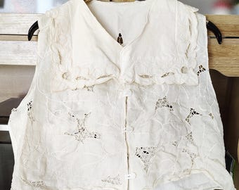 Peut inclure: Un gilet sans manches de couleur crème orné de broderies florales ajourées sur tout le tissu. Le vêtement présente un large col festonné et une fermeture frontale composée de trois petits boutons blancs assortis.