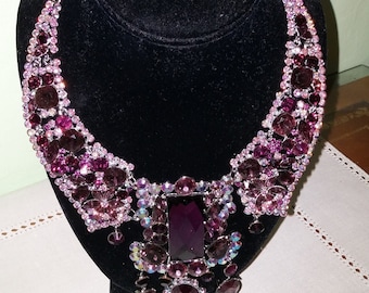 May include: A striking purple necklace and earring set, presented on a black bust. The necklace has a large rectangular central stone, surrounded by many smaller, shimmering purple and iridescent crystals. The earrings mirror the necklace's design.
