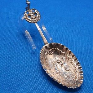 Puede incluir: Una cuchara de plata con un mango adornado con el rostro de una mujer y un diseño detallado en el cuenco. La cuchara está sobre un fondo azul.