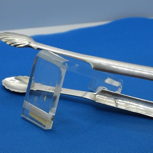 May include: Silver sugar tongs with a shell-shaped design on the ends. The tongs are resting on a blue surface.