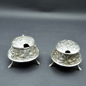 May include: Two silver sugar bowls with lids, each with intricate floral designs and three legs. The bowls are on a black surface.
