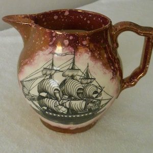 Puede incluir: Una jarra de cerámica roja y blanca con una imagen en blanco y negro de un barco de vela. La jarra tiene un esmalte moteado.