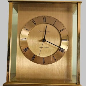 May include: A rectangular mantel clock with a gold-toned metal frame and glass panels. The clock face is circular with a brushed gold finish, black Roman numerals, and black hands. The text "Quartzmatic" and "SETH THOMAS" are printed on the face.
