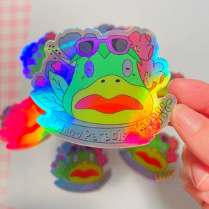 May include: Holographic sticker featuring a cartoon frog wearing sunglasses and a flower crown. The frog has a red mouth and is surrounded by flowers and leaves. The sticker says "Island Paradise" in a banner.