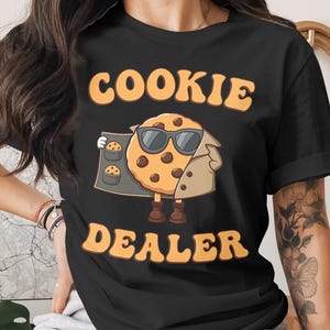 May include: Black t-shirt featuring the text "COOKIE DEALER" in orange. The design includes a cartoon cookie character wearing sunglasses and a trench coat, holding a tray of cookies.
