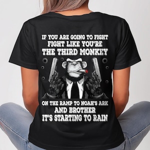 May include: Black t-shirt featuring a graphic of a monkey in a suit holding two guns and smoking a cigar. Text on the shirt reads "IF YOU ARE GOING TO FIGHT FIGHT LIKE YOU'RE THE THIRD MONKEY...IT'S STARTING TO RAIN."