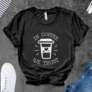 Può includere: Maglietta nera con testo bianco che recita "In Coffee We Trust" e un'immagine di una tazza di caffè con un cuore.