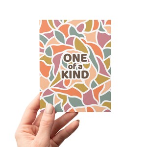 May include: A greeting card with a colorful abstract pattern in shades of pink, orange, yellow, and green. The card has the text "ONE of a KIND" in brown letters.