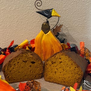May include: Two halves of a golden-brown pumpkin bread loaf positioned in front of a decorative wooden bird figurine with a yellow body and black head, surrounded by orange and black fabric scraps featuring spiderweb patterns.