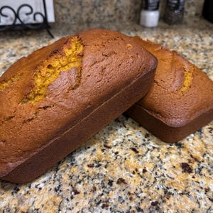 May include: Two rectangular loaves of golden-brown baked bread. Each loaf features a smooth, slightly domed top with a visible crack running lengthwise down the center, revealing a warm, orange-toned interior crumb texture.
