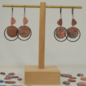 May include: Two pairs of dangle earrings with a circle and triangle design. The earrings are made of polymer clay with a marbled pattern in shades of pink, orange, blue, and green. The earrings are hanging from a wooden stand.