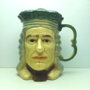 Puede incluir: Una taza de cerámica con forma de cabeza humana, con una corona verde y un cuello marrón. La taza tiene un asa y está pintada con un estilo realista.