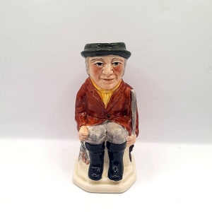May include: A ceramic figurine of a man wearing a brown jacket, green hat, and black boots. He is holding a gun and sitting on a white base.