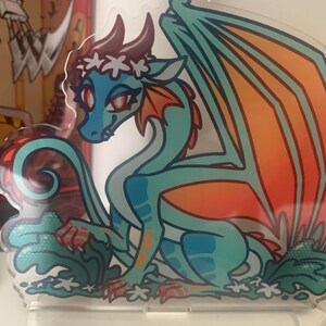 May include: A teal and orange dragon with a white flower crown, sitting on a white background with white flowers and green leaves. The dragon has red eyes and a pink nose.