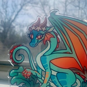 May include: A teal and orange dragon with a white flower crown, sitting on a green leafy base with white flowers. The dragon has a curled tail and orange wings.