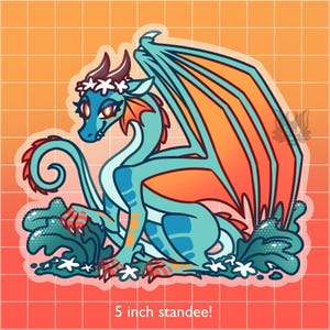 May include: A teal and orange cartoon dragon with a white flower crown and a curled tail. The dragon is sitting on a bed of green leaves with white flowers. The image is labeled "5 inch standee!"