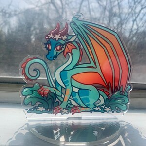 May include: A teal and orange dragon with a white flower crown sits on a clear acrylic stand. The dragon has a green tail and wings. The stand has a clear base and a textured surface.