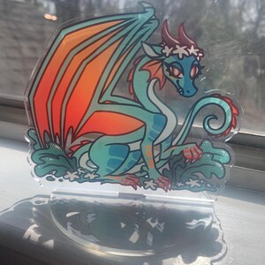 May include: A teal and orange dragon with a white flower crown, sitting on a clear acrylic stand with a green and white floral design.